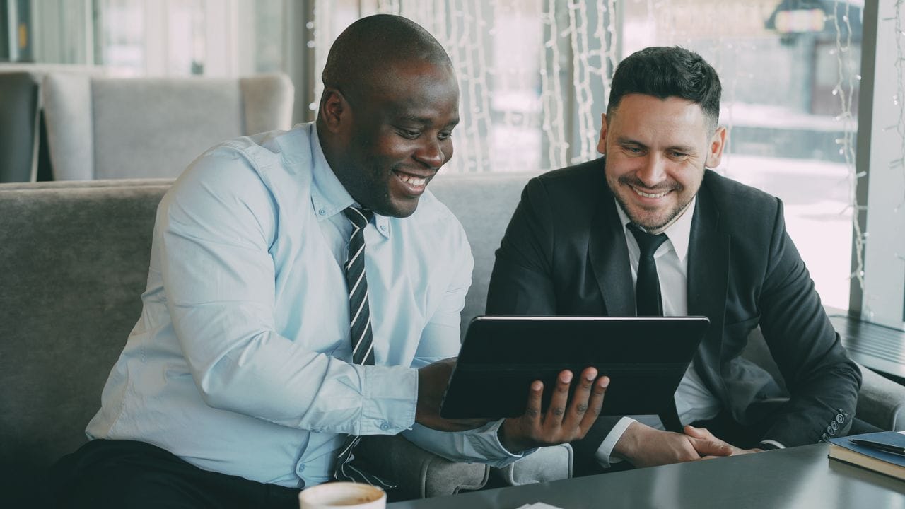 Offerings Two businessmen happily collaborating on a digital tablet in a modern cafe setting.