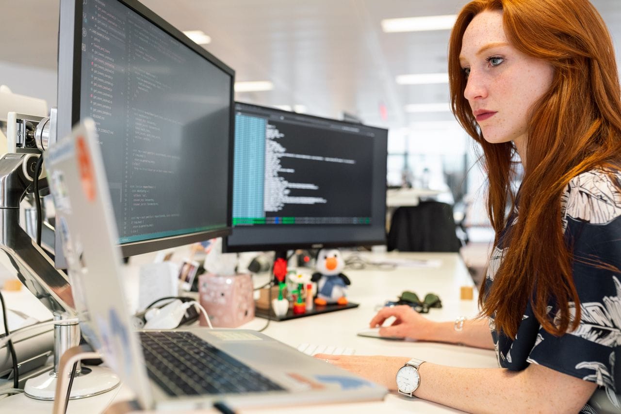 Offerings A focused female software engineer coding on dual monitors in a modern office.